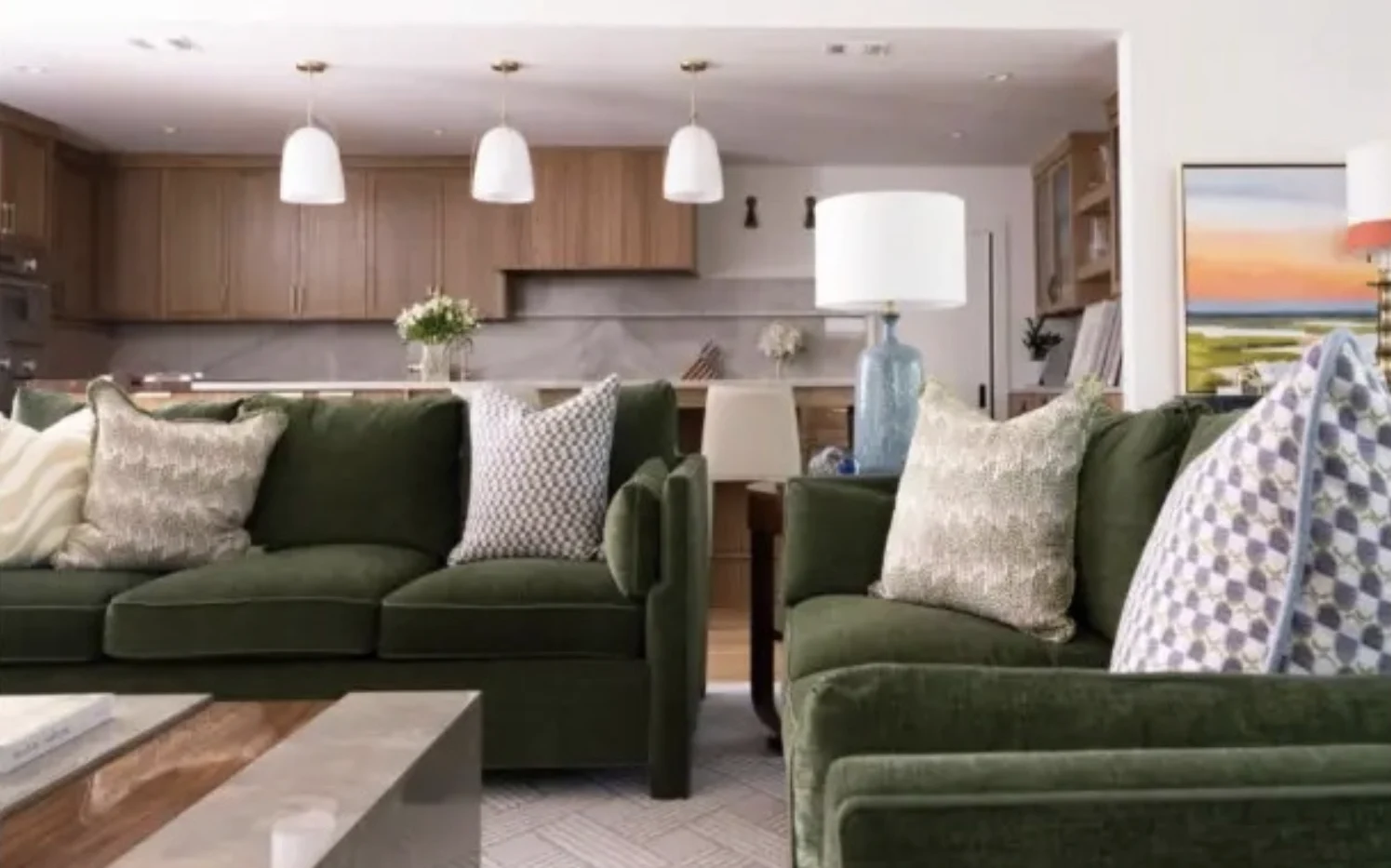 Living room with green velvet sofas, patterned pillows, and an open walnut kitchen beyond